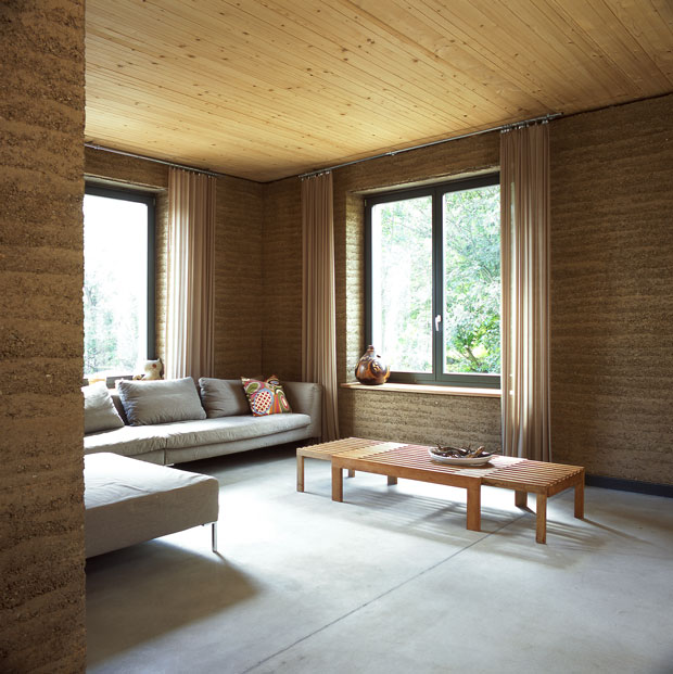 Wohnraum mit Sofa und zwei Fenstern. Die Decke ist aus hellem Holz, die Wände sind mit Lehm verputzt. Wohnraum mit Sofa und zwei Fenstern. Die Decke ist aus hellem Holz, die Wände sind mit Lehm verputzt.