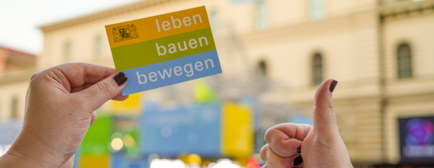 Im Vordergrund hält eine Hand ein Kärtchen mit Text "leben bauen bewegen". Eine andere Hand hält einen ausgestreckten Daumen in die Kamera. Im Hintergrund ist unscharf der IAA-Messestand des StMB zu erkennen. Im Vordergrund hält eine Hand ein Kärtchen mit Text "leben bauen bewegen". Eine andere Hand hält einen ausgestreckten Daumen in die Kamera. Im Hintergrund ist unscharf der IAA-Messestand des StMB zu erkennen.