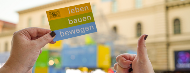 Im Vordergrund hält eine Hand ein Kärtchen mit Text "leben bauen bewegen". Eine andere Hand hält einen ausgestreckten Daumen in die Kamera. Im Hintergrund ist unscharf der IAA-Messestand des StMB zu erkennen.