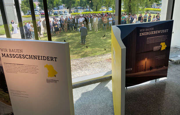 Zwei Stelen der Wanderausstellung stehen in einem Ausstellungsraum. Durch Glasfenster sind viele Personen zu sehen, die auf einer Wiese stehen und einem Mann, der eine Rede hält, zuhören.