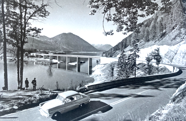 Schwarz-weiß-Foto mit einem Auto aus den 50er-Jahren im Vordergrund, das über eine geschwungene Straße fährt. Die Straße führt an einem See vorbei und als Brücke mit sieben Pfeilern über den See. Im Hintergrund sind Berge zu sehen. Schwarz-weiß-Foto mit einem Auto aus den 50er-Jahren im Vordergrund, das über eine geschwungene Straße fährt. Die Straße führt an einem See vorbei und als Brücke mit sieben Pfeilern über den See. Im Hintergrund sind Berge zu sehen.