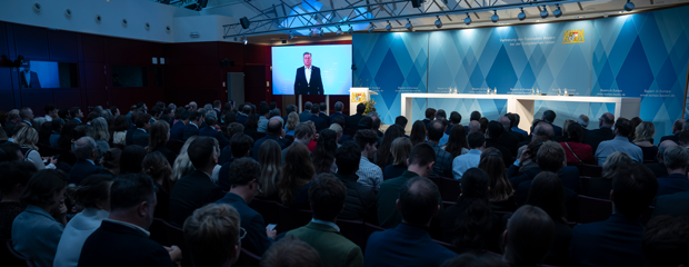 Blick in einen vollbesetzten Saal. Auf einer Leinwand ist Staatsminister Christian Bernreiter zu sehen, der ein Grußwort hält. Blick in einen vollbesetzten Saal. Auf einer Leinwand ist Staatsminister Christian Bernreiter zu sehen, der ein Grußwort hält.