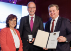 Verkehrsminister Christian Bernreiter überreicht eine Urkunde und eine Medaille an einen Mann. Daneben steht die Laudatorin.