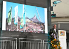 Hier ist ein Herr auf einer Bühne am Rednerpult zu sehen. Im Hintergrund auf einer Leinwand ein Bild von den Olympischen Spielen 1972.