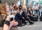 Menschen sitzen und stehen um die Bühne im Foyer und hören einem Fachvortrag zu.