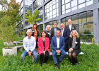 Gruppenfoto der Beteiligten am Impulsformat "Lebenswerte Stadt der Zukunft". Die Beteiligten sitzen und stehen vor einem Gebäude. Im Hintergrund ist viel Grün (Wiese, Bäume) zu sehen.