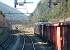 Auf diesem Bild ist ein Güterzug von der Seite zu sehen. Im Hintergrund sind Berge und Bäume zu sehen. Neben dem Zug weitere Gleise und Oberleitungen. 