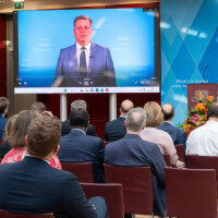 Verkehrsminister Christian Bernreiter ist auf einer Leinwand bei einer Videobotschaft zu sehen. Die Leinwand ist vor Publikum aufgebaut, das in einem Veranstaltungsraum sitzt.