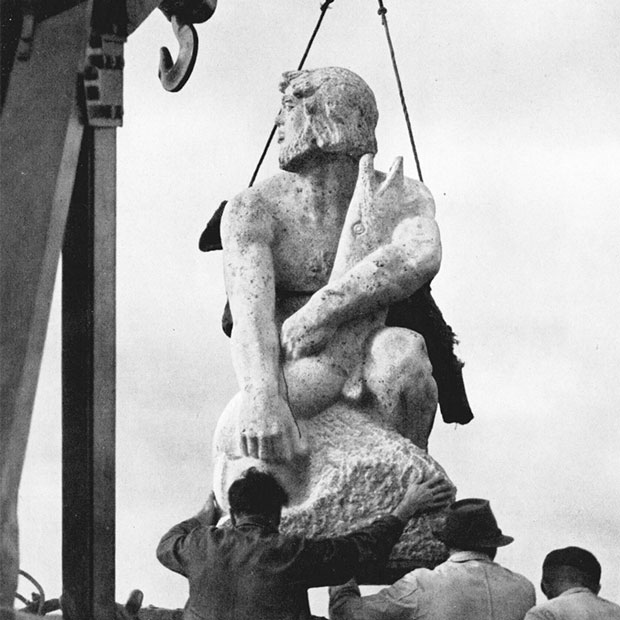 Eine große Statue aus Stein (Ein Flussgott, der einen großen Fisch im Arm hält) wird von einem Kran abgeladen. Drei Männer begleiten die Lieferung Eine große Statue aus Stein (Ein Flussgott, der einen großen Fisch im Arm hält) wird von einem Kran abgeladen. Drei Männer begleiten die Lieferung
