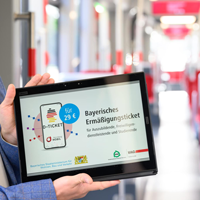 Ein Tablet-Bildschirm, der von zwei Händen ins Bild gehalten wird. Auf dem Bildschirm ist ein Handy mit dem Logo des Deutschlandticket zu sehen und der Text "Bayerisches Ermäßigungsticket für Auszubildende, Freiwilligendienstleistende und Studierende". Im Hintergrund ist unscharf das Innere einer S-Bahn zu erkennen.