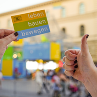 Im Vordergrund hält eine Hand ein Kärtchen mit Text "leben bauen bewegen" in die Kamera, eine andere Hand zeigt einen nach oben gestreckten Daumen. Im Hintergrund ist unscharf der Messestand des StMB zu erkennen.