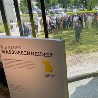 Zwei Stelen der Wanderausstellung stehen in einem Ausstellungsraum. Durch Glasfenster sind viele Personen zu sehen, die auf einer Wiese stehen und einem Mann, der eine Rede hält, zuhören.