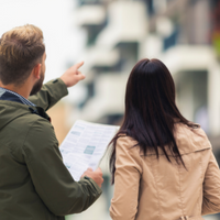 Ein Mann und eine Frau stehen vor einem Wohngebäude. Der Mann hält in der Hand Blatt mit Wohnungsanzeigen und deutet auf das Gebäude, das unscharf zu sehen ist. 