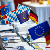Verschiedene kleine Flaggen, u.a. die europäische, deutsche und bayerische Flagge, stehen als Dekoration auf einem Tisch, auf dem verschiedene Flyer und Broschüren ausgelegt sind