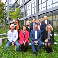 Gruppenfoto der Beteiligten am Impulsformat "Lebenswerte Stadt der Zukunft". Die Beteiligten sitzen und stehen vor einem Gebäude. Im Hintergrund ist viel Grün (Wiese, Bäume) zu sehen.