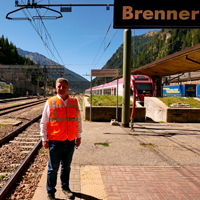 Hier steht Herr Staatsminister Christian Bernreiter an einem Gleis am Brenner. Der Himmel ist blau. Im Hintergrund sind Gleise und ein roter Zug zu sehen. 