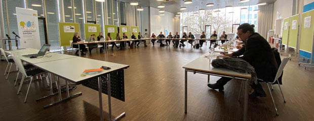 In einem großen Besprechungsraum sitzen an einem sehr großen, U-förmigen Besprechungstisch mehrere Frauen und Männer. Im Hintergrund sind Rollups und Tafeln zu sehen. In einem großen Besprechungsraum sitzen an einem sehr großen, U-förmigen Besprechungstisch mehrere Frauen und Männer. Im Hintergrund sind Rollups und Tafeln zu sehen.