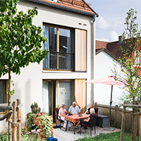 Bewohnerinnen und Bewohner sitzen auf einer Terrasse vor einem Wohngebäude Bewohnerinnen und Bewohner sitzen auf einer Terrasse vor einem Wohngebäude