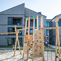 Wohngebäude mit Holzfassade. Vor dem Gebäude ein Spielplatz-Klettergerüst mit einem Kind Wohngebäude mit Holzfassade. Vor dem Gebäude ein Spielplatz-Klettergerüst mit einem Kind